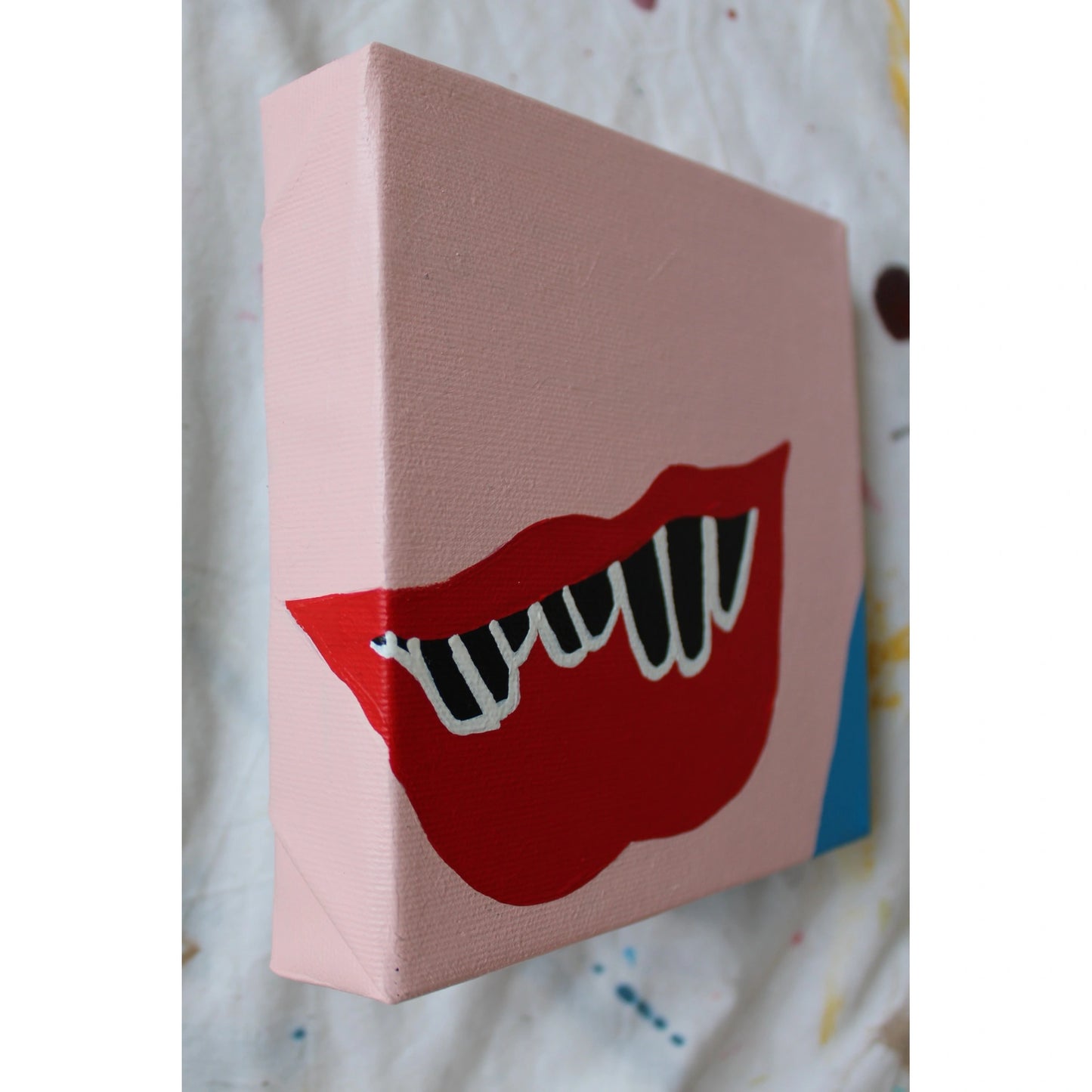 "Side view of a small pink canvas featuring a red open mouth with sharp white fangs and black gaps, painted in minimalist pop art style, part of PLAY ME. BOY. by Liya Ella Raspberry."