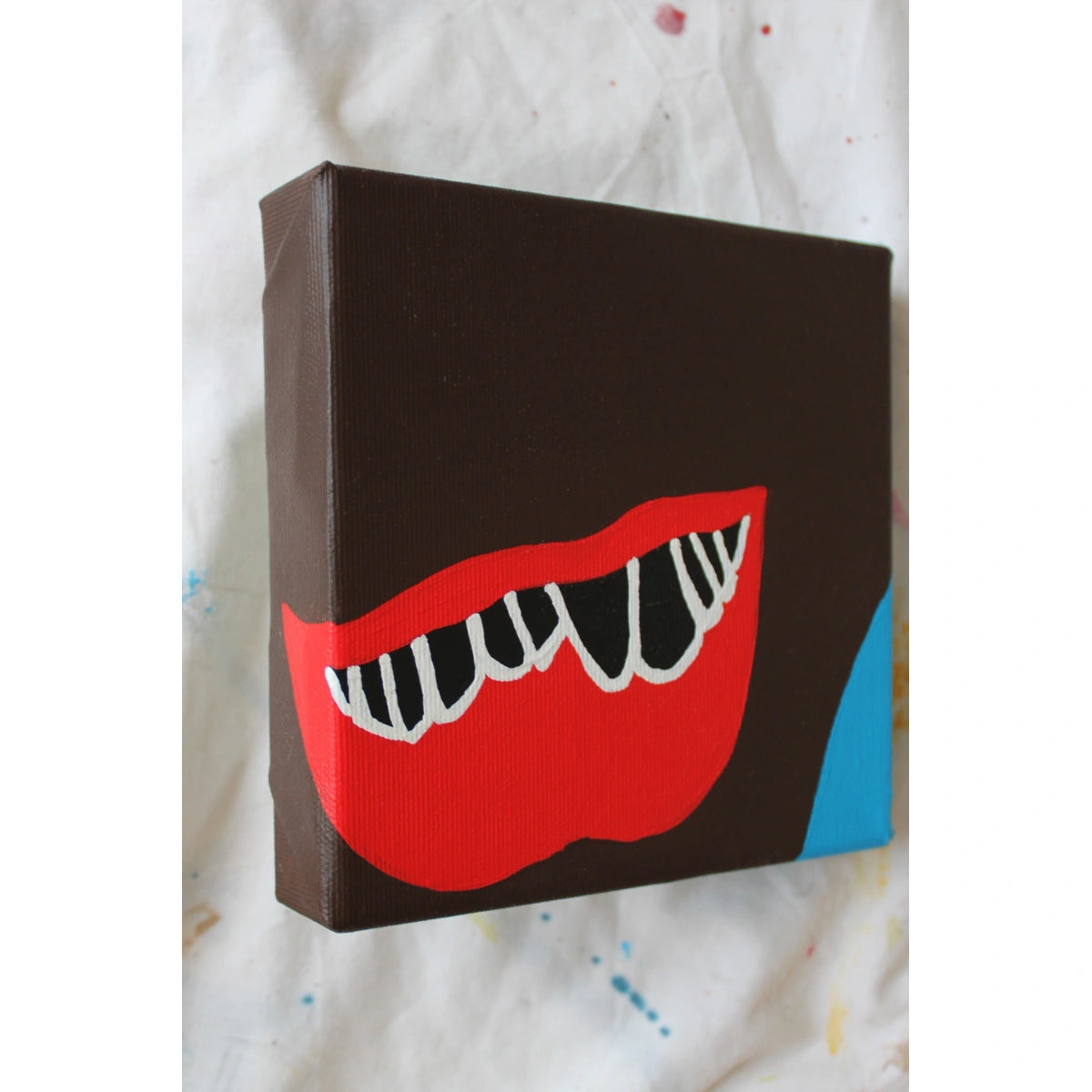 "Side view of a contemporary red lips painting with expressive white teeth on dark brown canvas and blue corner — acrylic artwork by Liya Ella Raspberry, part of the PLAY ME. BOY. collection, 2025."