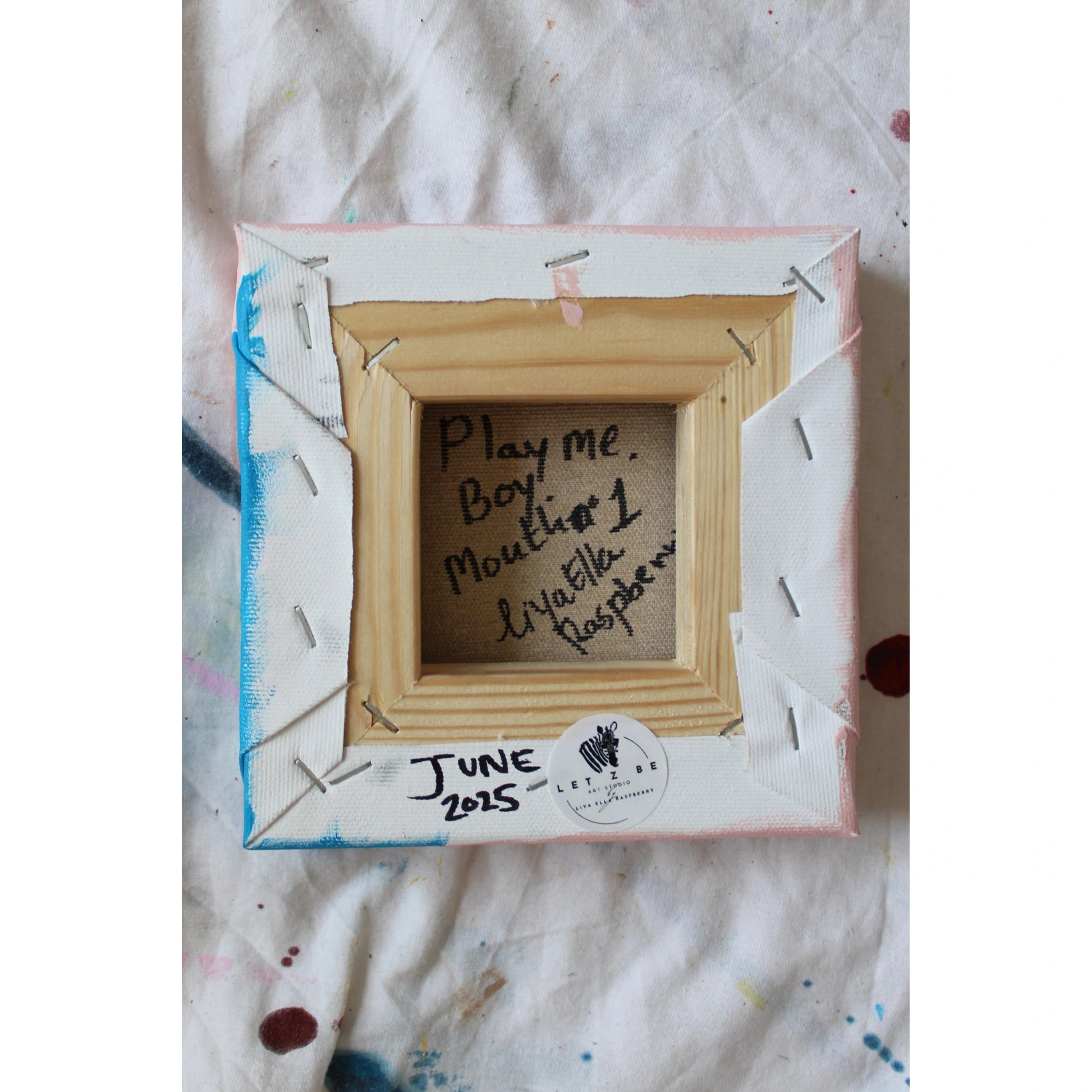 "Back of a signed mini canvas by Liya Ella Raspberry from the PLAY ME. BOY. series, titled 'Mouth #1,' dated June 2025, with Let Z Be logo sticker."